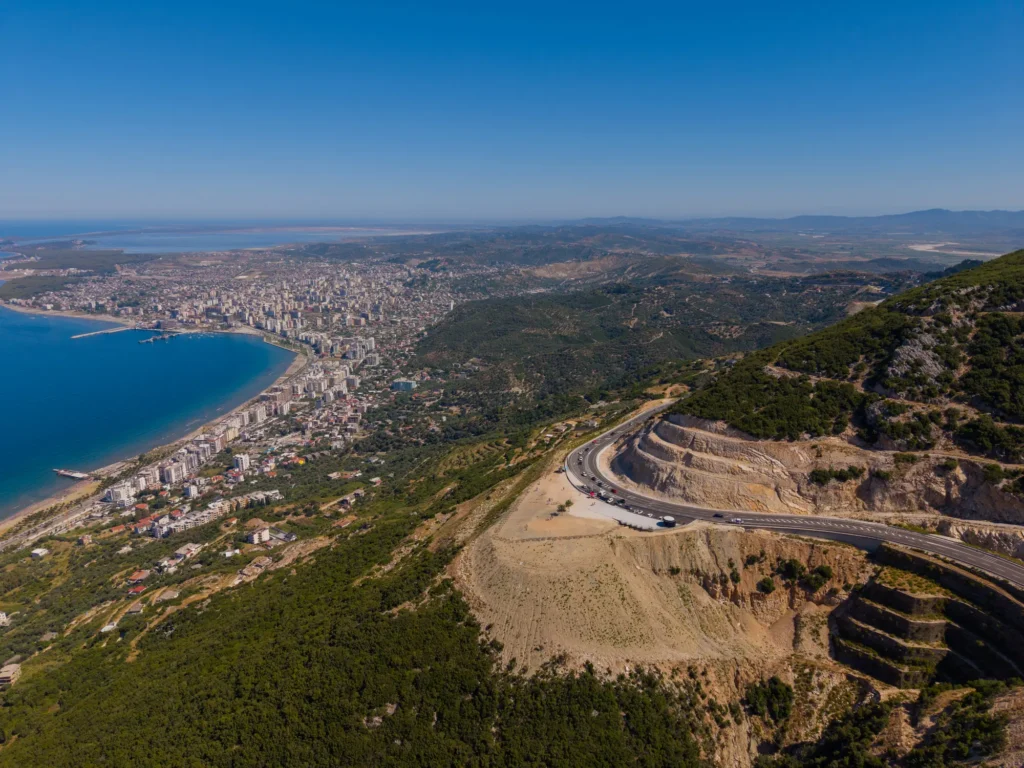 Aerial view of Vlorë, Albania, showing the coastal city along the bay with a winding mountain road in the foreground