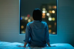 A woman sits alone on a bed, facing a window overlooking an unfamiliar city at night