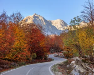 Winding autumn road through the Albanian Alps near the Valbona River, with snow-capped peaks in the background